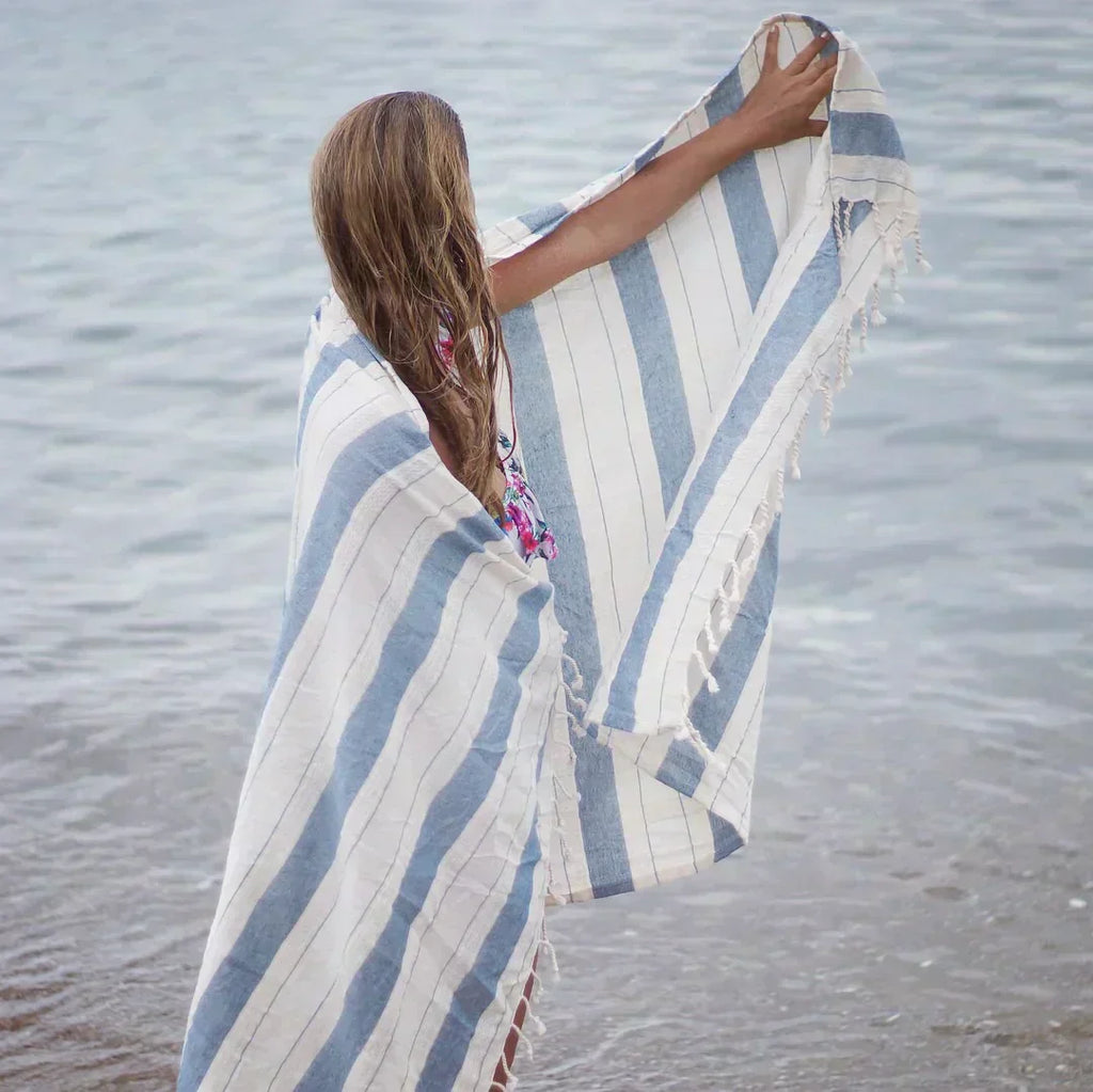 En jente med et stripete lyseblått badehåndkle rundt seg som ser utover havet.